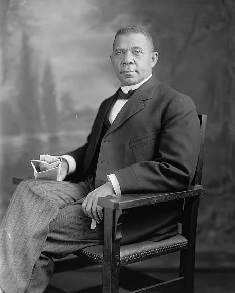 Black and white portrait of Booker T. Washington. Washington is seated in a chair with his right leg crossing his left. In his left hand he hold a folded piece of paper. Washington wears a dark suit coat, a dark vest, and a white shirt with a dark bow tie. He wears grey pinstriped trousers.